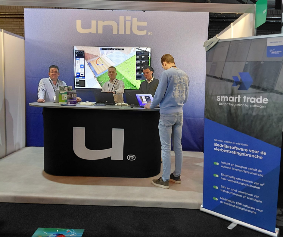 Beursstand van Unlit met drie medewerkers achter een balie en een bezoeker die een laptop bekijkt, naast een Smart Trade banner over bedrijfssoftware voor de sierbestratingsbranche.