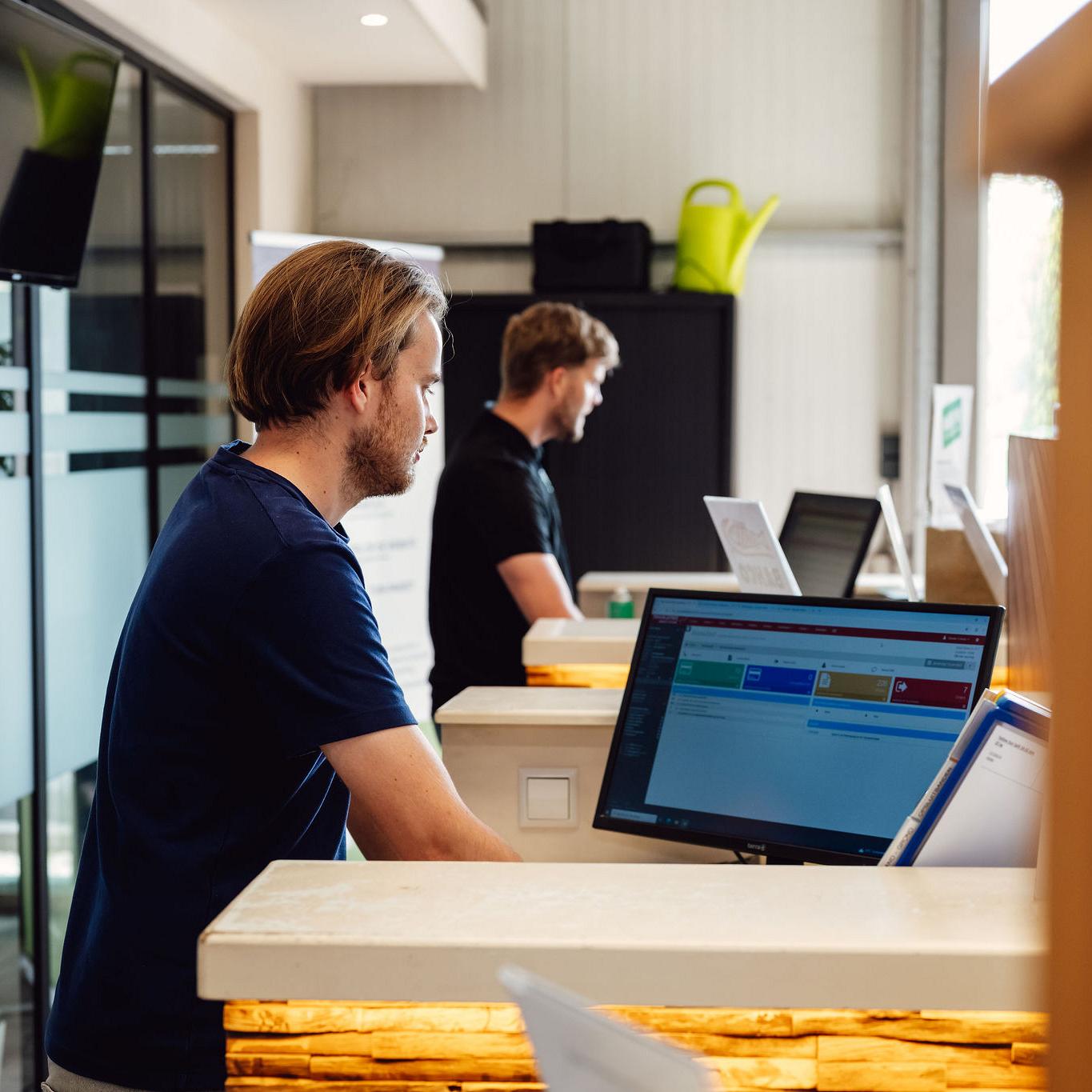 Twee medewerkers werken achter balies met computers in een moderne kantoorruimte; op het scherm is Smart Trade-software zichtbaar.