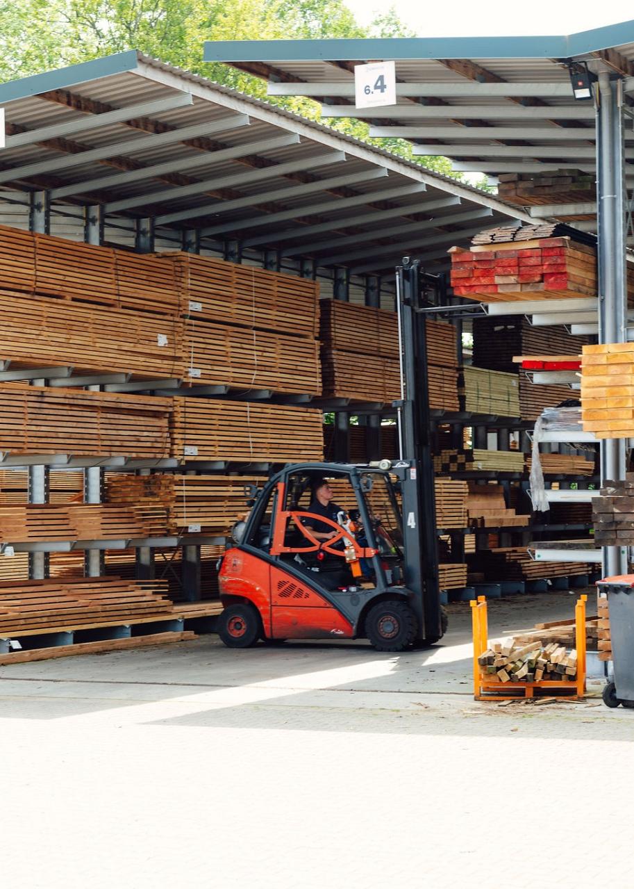 Buitenopslag met houten balken op metalen rekken, waar een heftruck bezig is met het verplaatsen van materialen onder een overkapping.