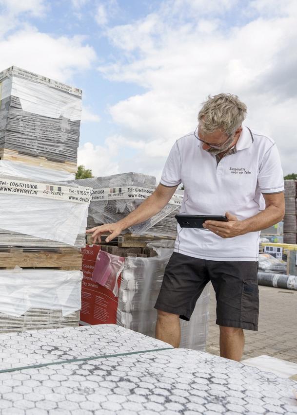 Man in witte polo controleert buiten een voorraad pallets met natuursteen of tegels, met behulp van een tablet en omringd door gestapelde verpakte materialen.