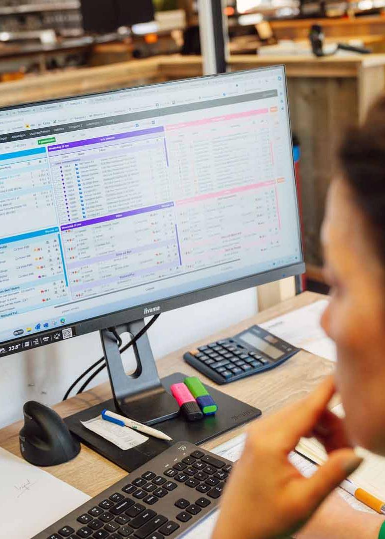 Persoon werkt aan bureau met twee monitoren met spreadsheets en grafieken, notitieblok en pen voorhanden.