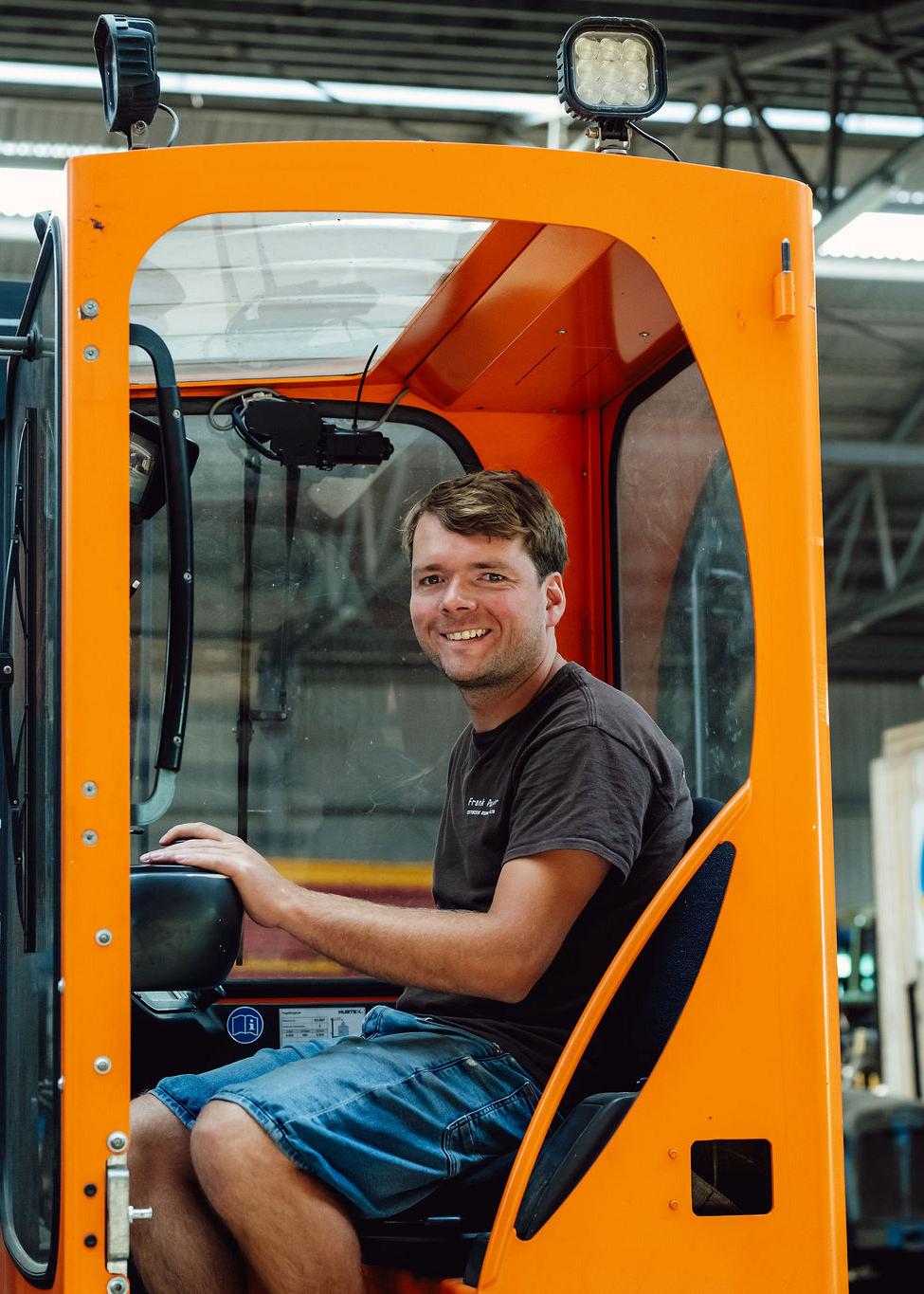 Lachende medewerker zit in een oranje heftruck binnen een werkplaats, klaar om goederen te verplaatsen.