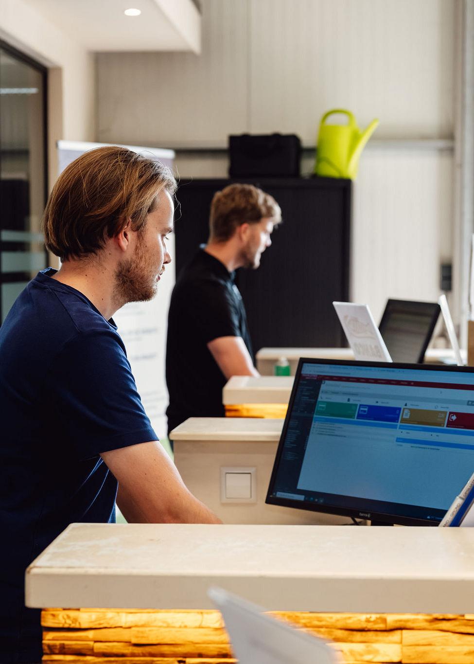 Twee medewerkers werken achter balies met computers in een moderne kantoorruimte; op het scherm is Smart Trade-software zichtbaar.