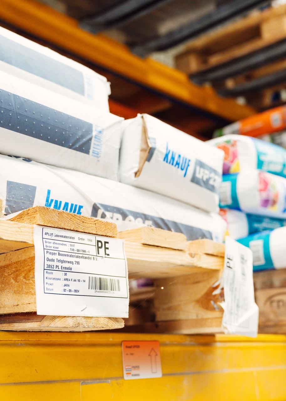 Close-up van pallets met bouwmaterialen gestapeld op stellingen in een magazijn, voorzien van labels met productinformatie.
