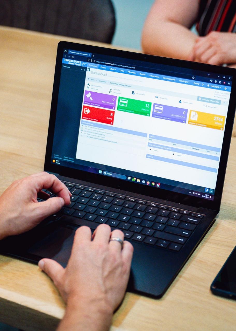 Persoon werkt op een laptop met een digitaal beheersysteem op het scherm, zittend aan een houten tafel met een smartphone en een andere persoon tegenover.