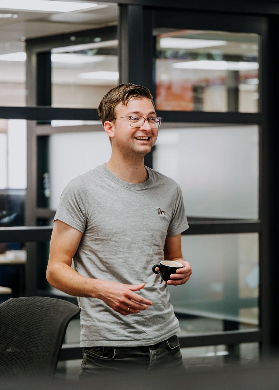Twee collega’s praten en lachen in een moderne kantoorruimte met glazen wanden; een van hen houdt een bril in zijn hand.