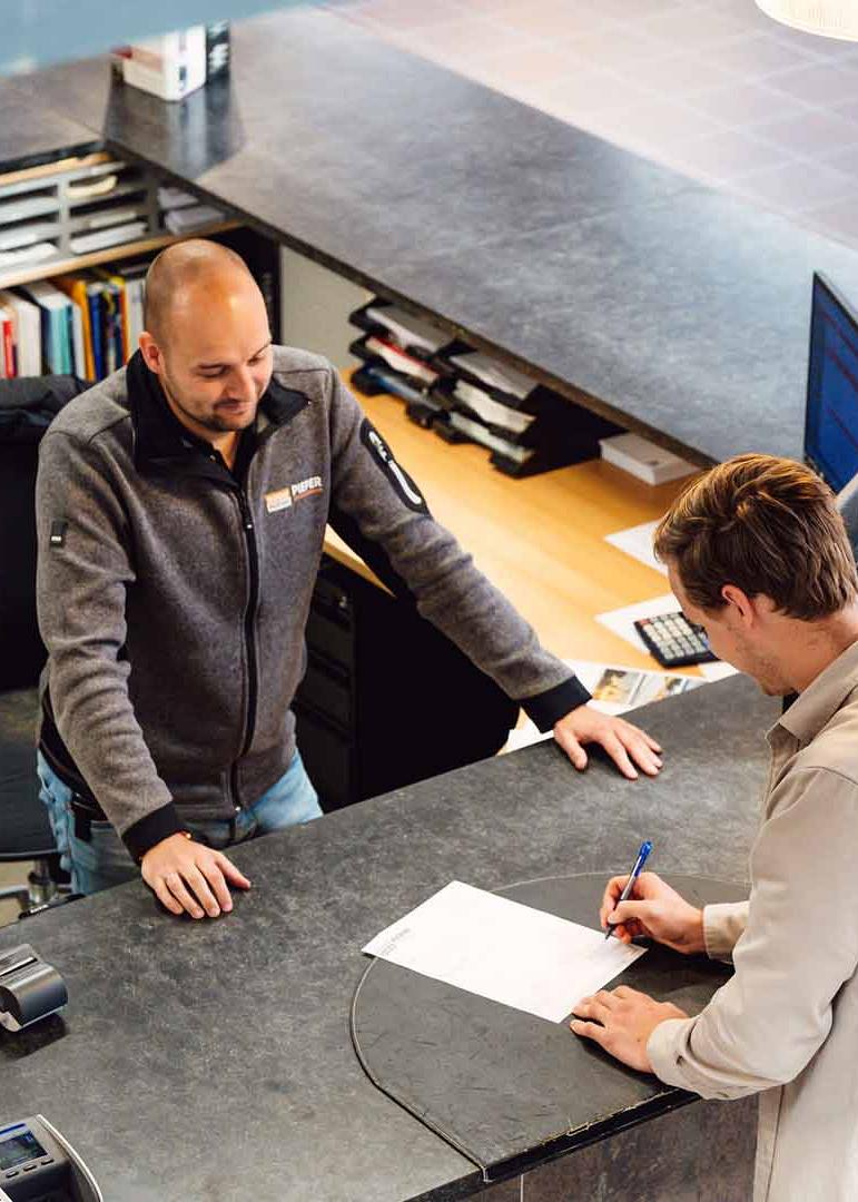 Man ondertekent een formulier aan een balie terwijl een medewerker naar hem kijkt.