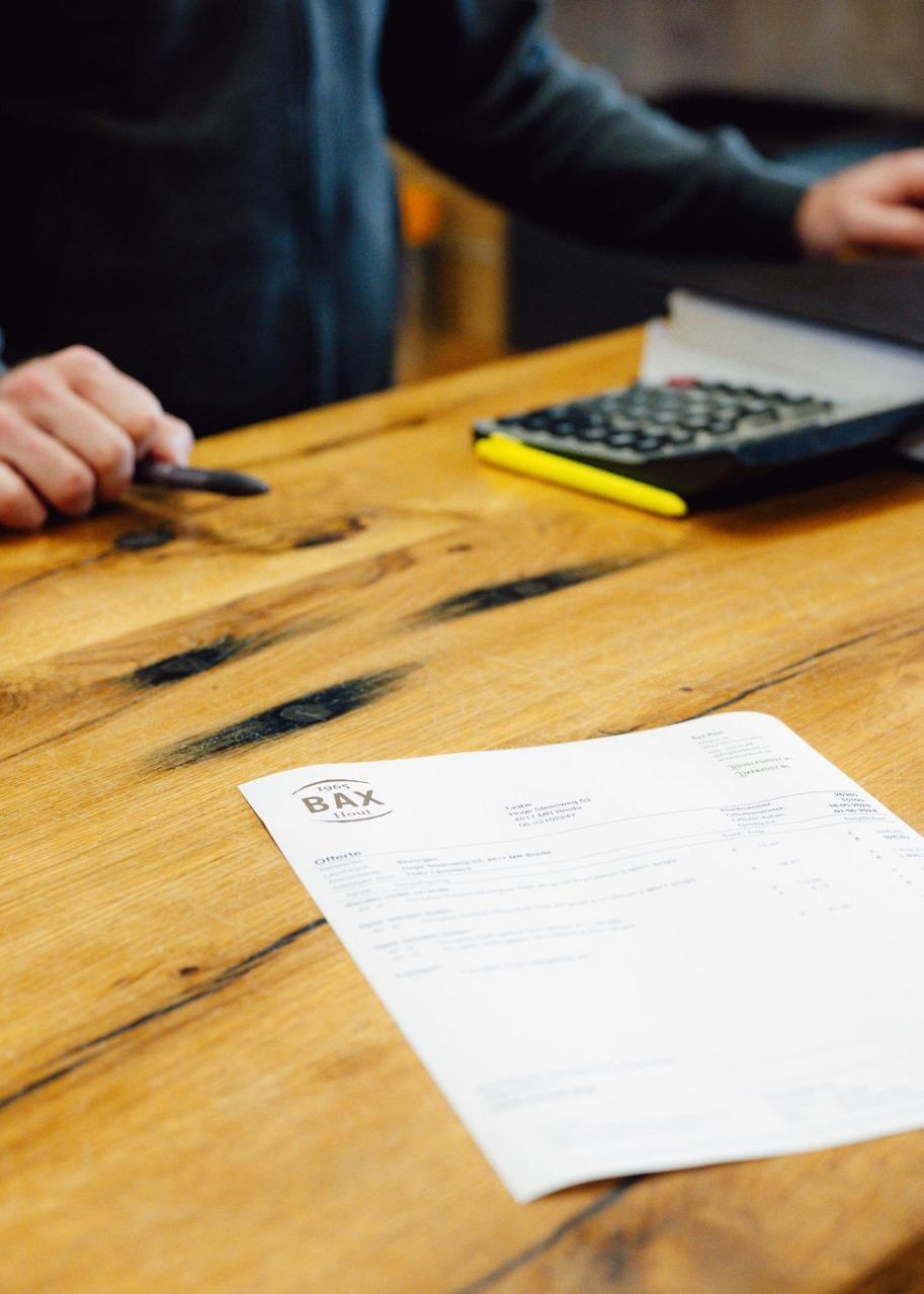 Offerte op houtentafel met pennenbak, rekenmachine, agenda en hand die pen vasthoudt.