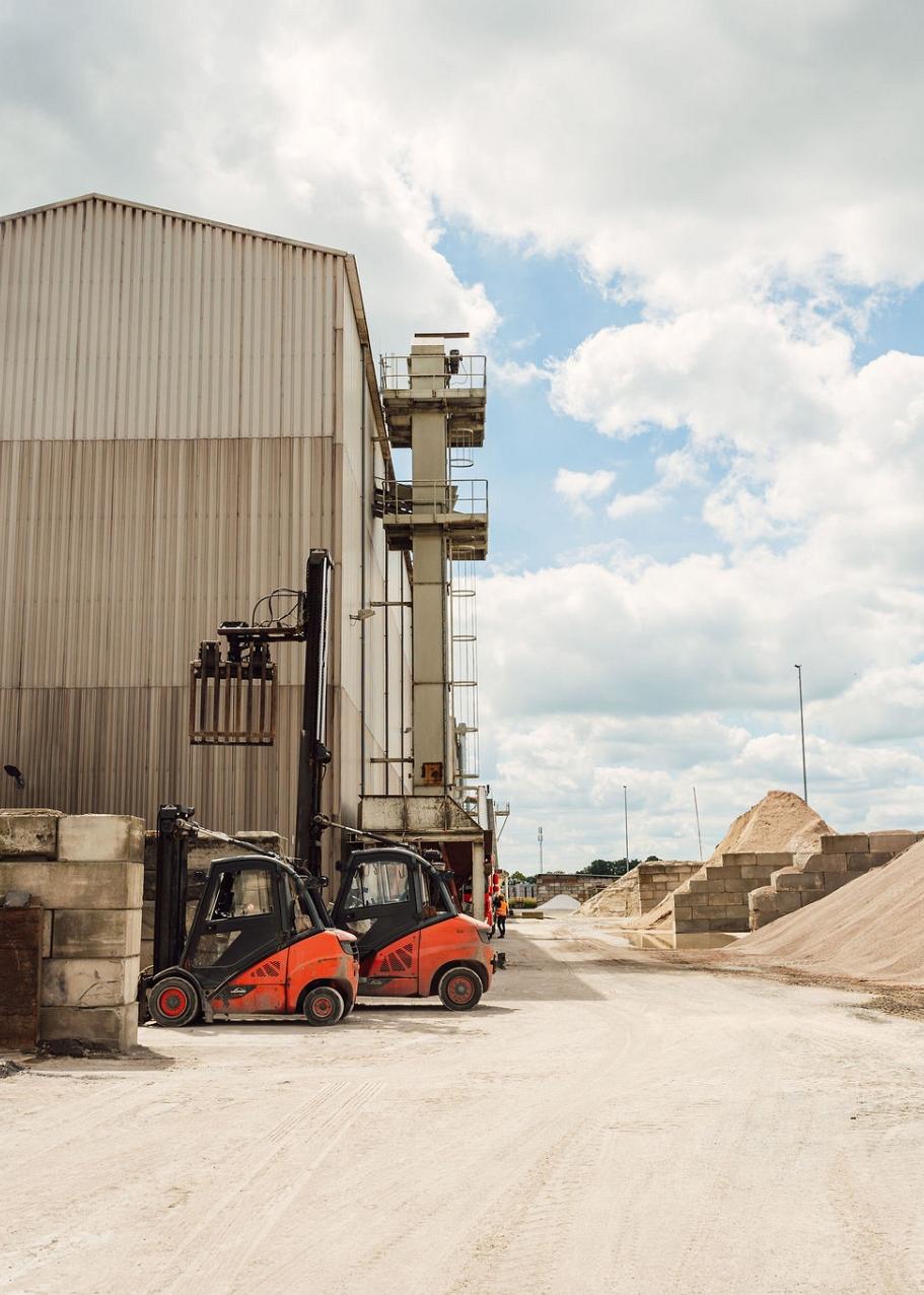 Een terrein met stapels betonblokken links, een groot gebouw rechtachterin en linksachterin een betonnen afscheiding met een grote berg zand.