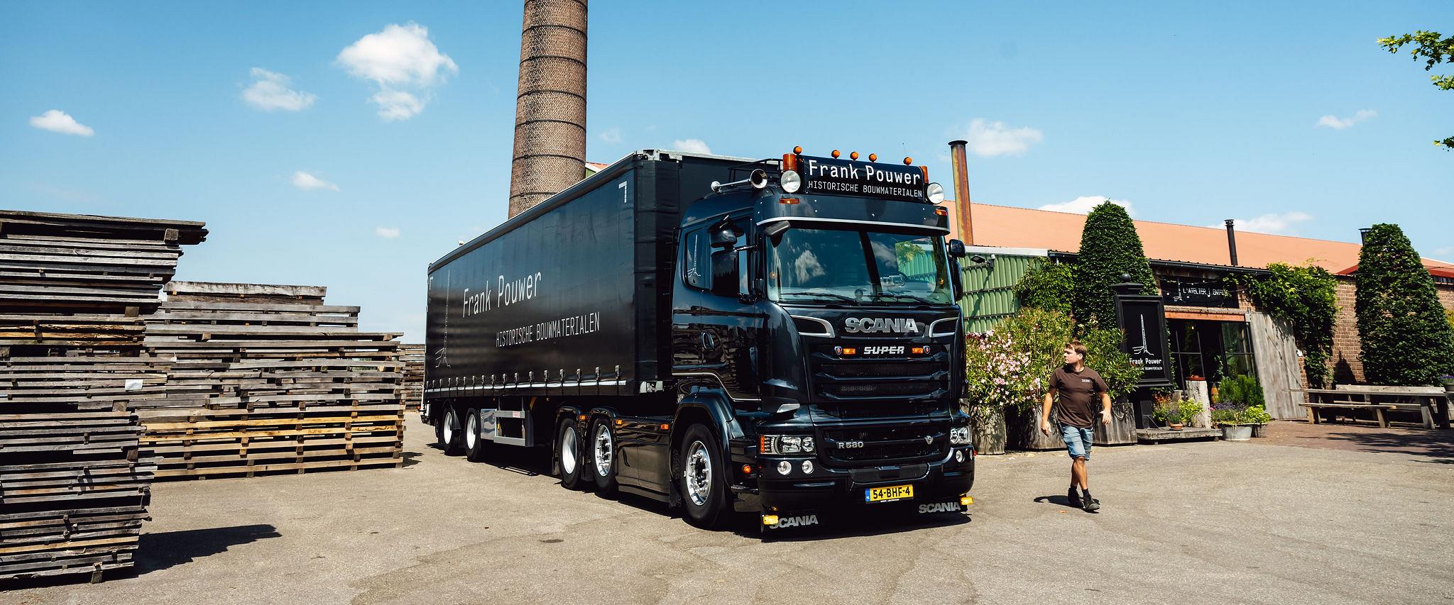 Zwarte vrachtwagen van een houthandel op een buitenterrein met stapels hout, een hoge schoorsteen en een medewerker in werkkleding.