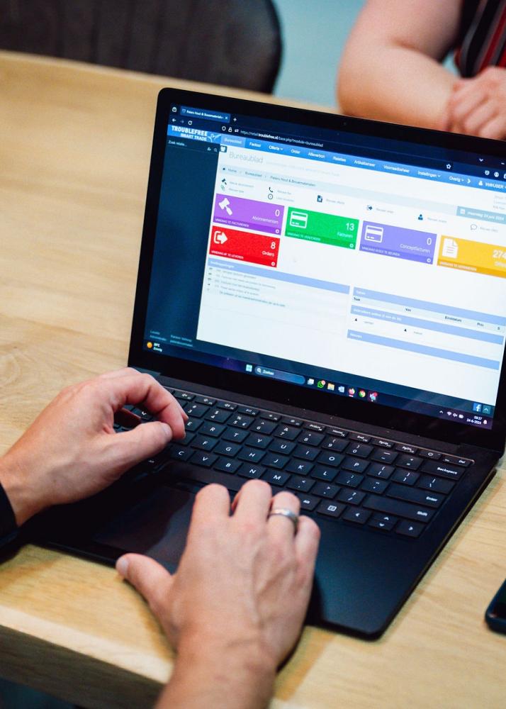 Persoon werkt op een laptop met een digitaal beheersysteem op het scherm, zittend aan een houten tafel met een smartphone en een andere persoon tegenover.