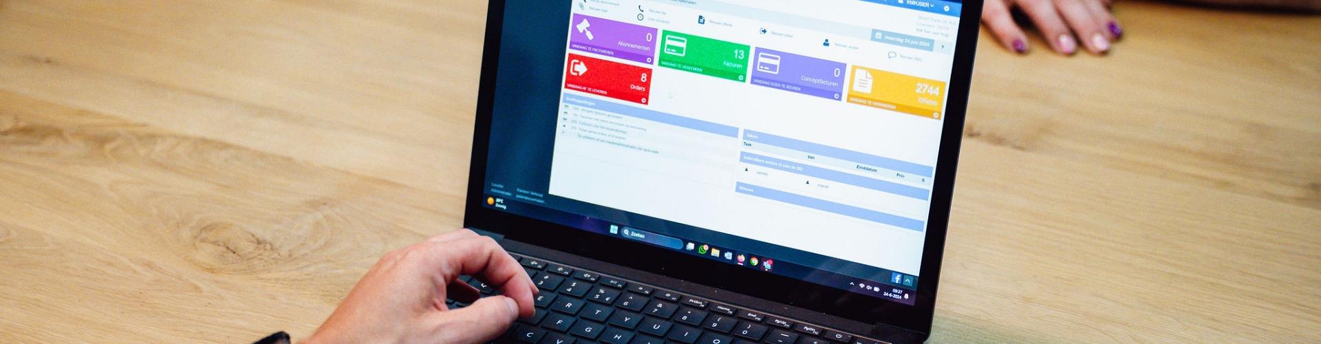 Persoon werkt op een laptop met een digitaal beheersysteem op het scherm, zittend aan een houten tafel met een smartphone en een andere persoon tegenover.