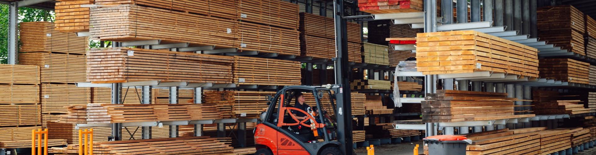 Buitenopslag met houten balken op metalen rekken, waar een heftruck bezig is met het verplaatsen van materialen onder een overkapping.