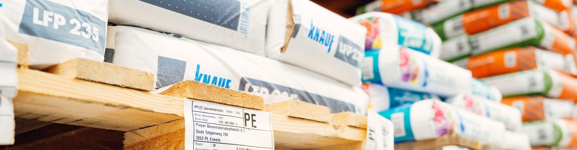 Close-up van pallets met bouwmaterialen gestapeld op stellingen in een magazijn, voorzien van labels met productinformatie.