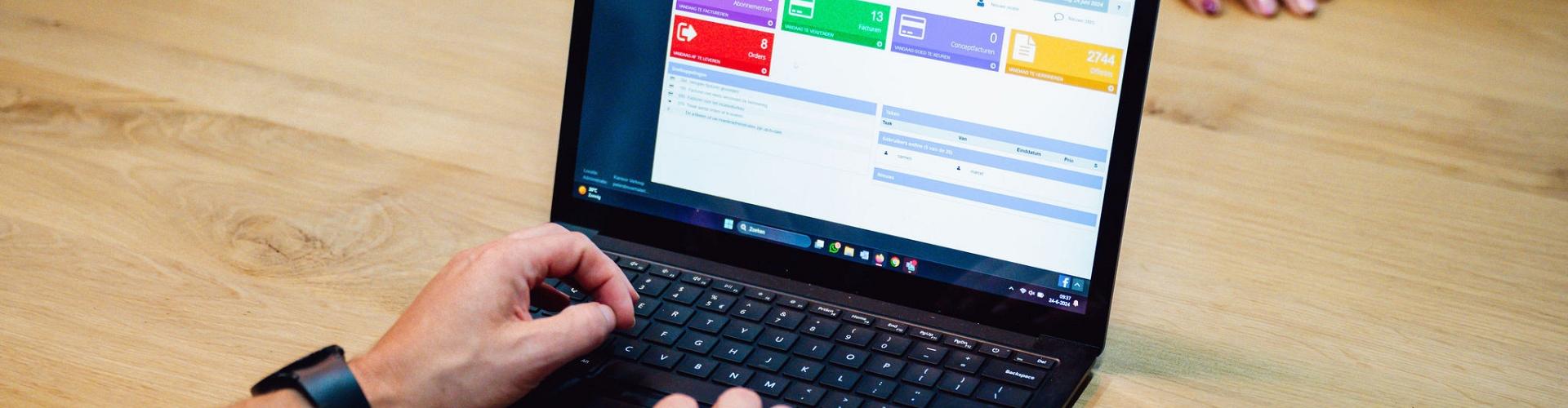 Persoon werkt op een laptop met een digitaal beheersysteem op het scherm, zittend aan een houten tafel met een smartphone en een andere persoon tegenover.