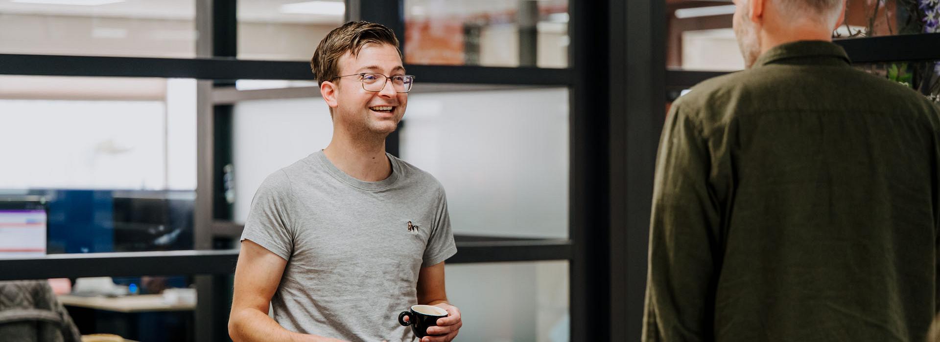 Twee collega’s praten en lachen in een moderne kantoorruimte met glazen wanden; een van hen houdt een bril in zijn hand.