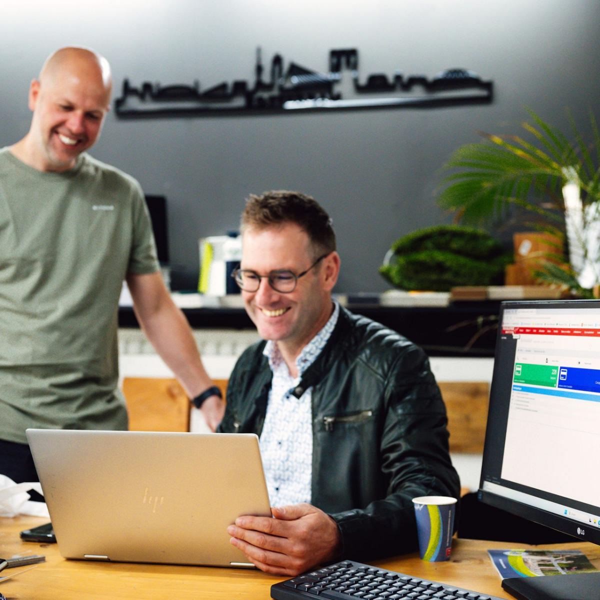 Twee lachende mannen, 1 staat en 1 zit achter een bureau. Een laptop of tafel.