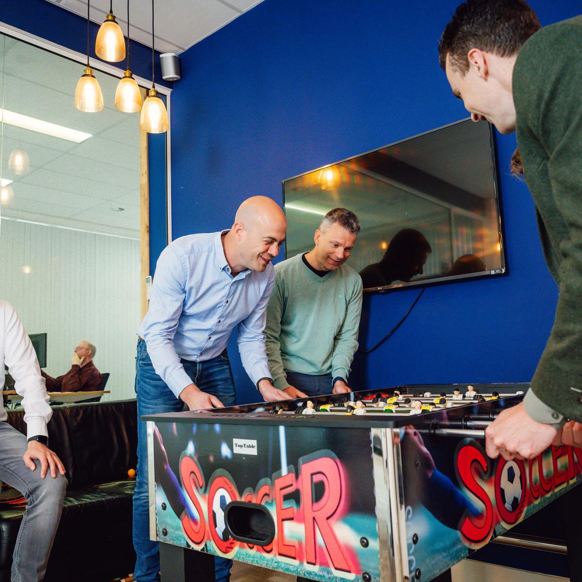 Vier directieleden lachen en spelen tafelvoetbal in een moderne kantoorruimte met een blauwe muur en glazen vergaderruimte op de achtergrond.