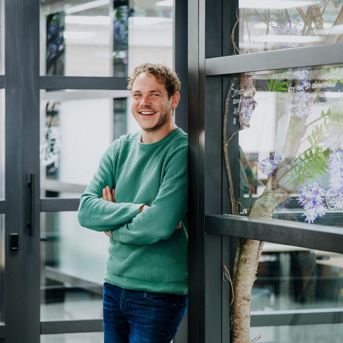 Lachende man in groene trui leunt ontspannen tegen een glazen wand in een moderne kantoorruimte met planten en bureaus op de achtergrond.