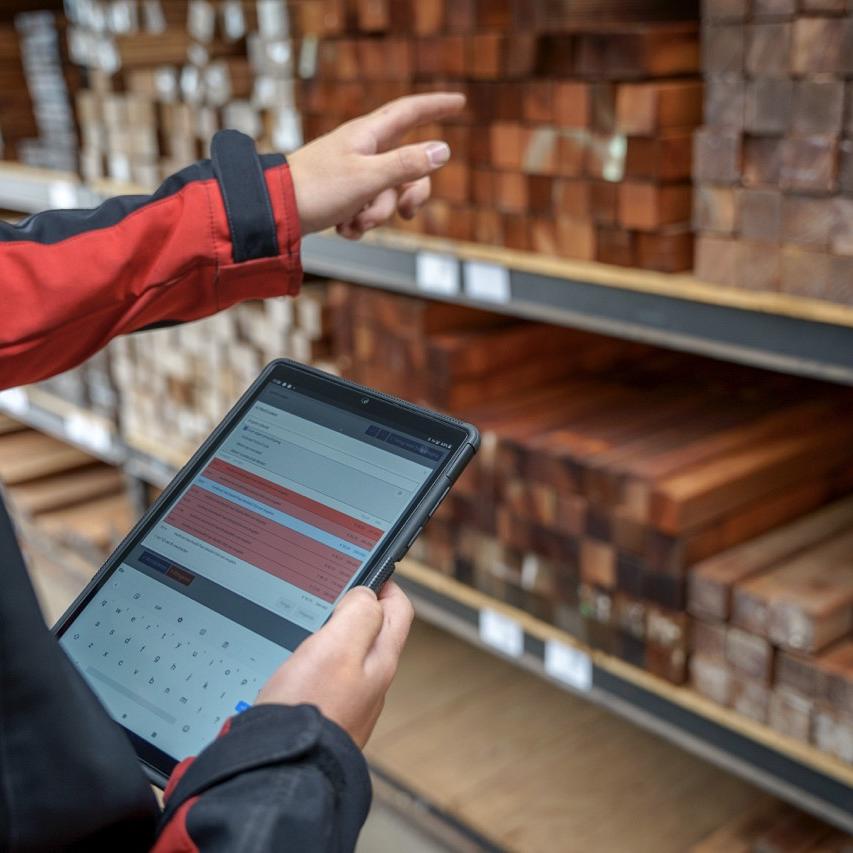 Handen die een tablet vasthouden met daarop Smart Trade software en de andere hand wijst naar een stelling met hout blokken. 