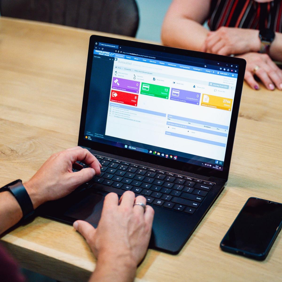 Laptop op tafel waar een man op aan het werk is met daarnaast een telefoon en aan de andere kant van de tafel zit een vrouw met haar handen over elkaar.