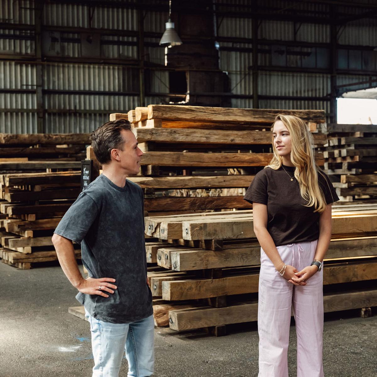 Twee mensen in gesprek in een loods met opgestapelde houten balken; ze staan tussen de stapels en lijken iets te bespreken over het hout.