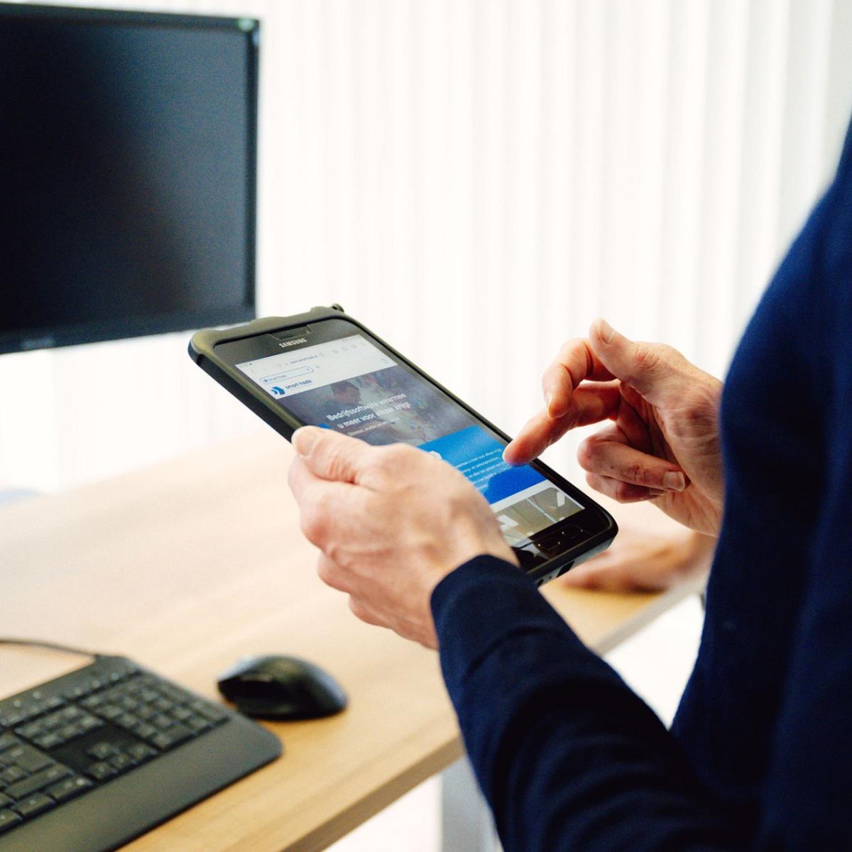 Persoon in donkerblauwe kleding bedient een tablet met de website van Smart Trade open, staand bij een bureau met een monitor, toetsenbord en muis.