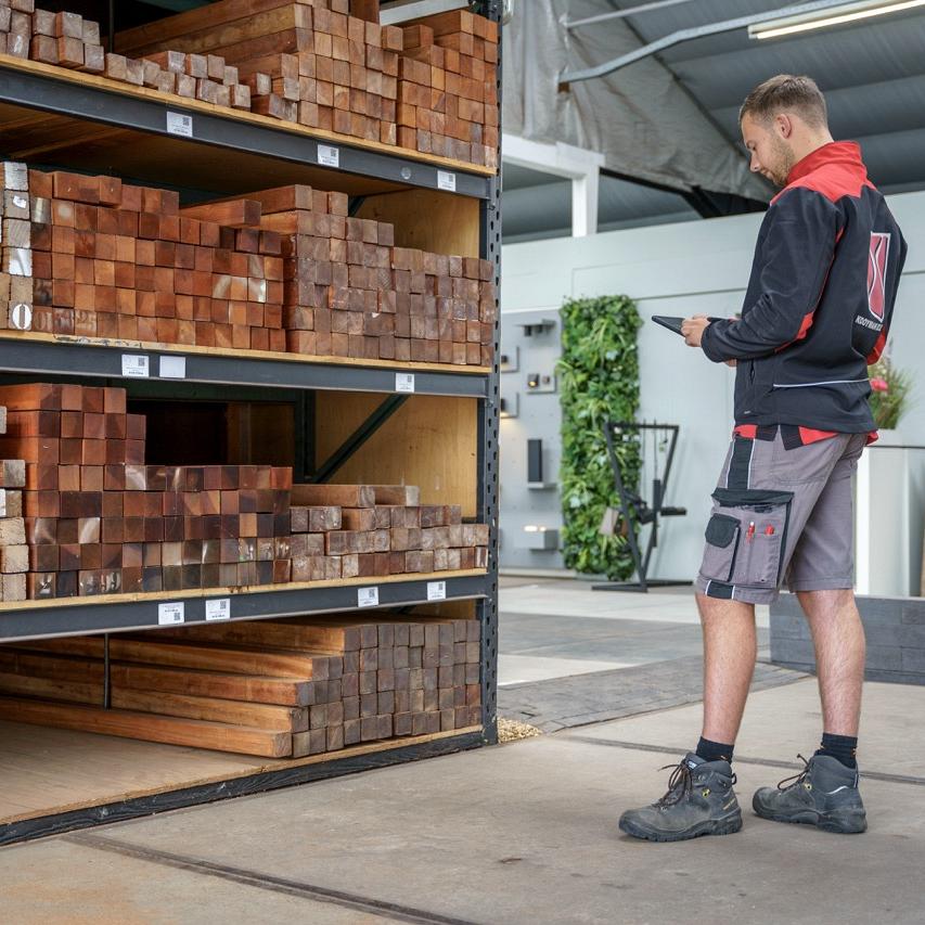 Medewerker in werkoutfit controleert de voorraad houten balken in een magazijn met een tablet, terwijl de planken netjes gestapeld liggen in metalen rekken.