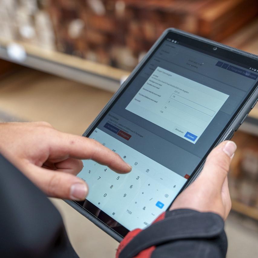 Persoon in werkoutfit gebruikt een tablet met een digitaal voorraadbeheersysteem, staand bij stellingen met houten balken in een magazijn.