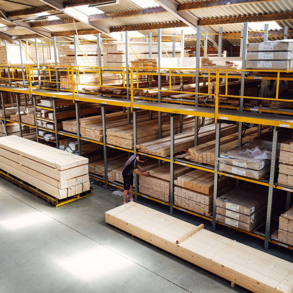 Ruim magazijn met hoge stellingen vol houten platen en balken, waar een medewerker bezig is met een grote stapel hout op de vloer.