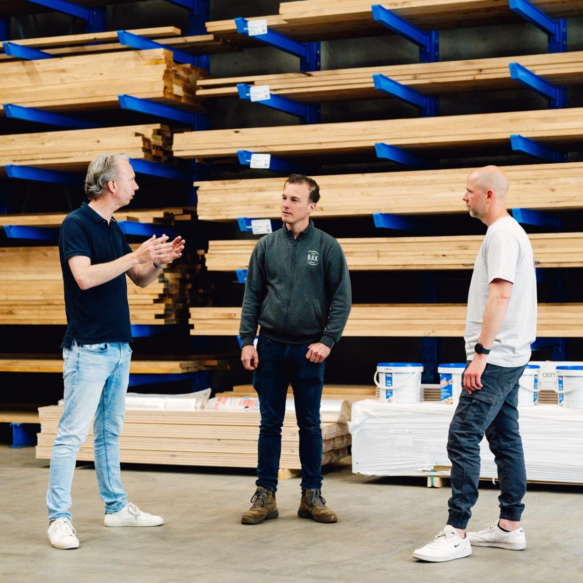 Drie mannen overleggen in een magazijn met lange houten balken op metalen stellingen, terwijl bouwmaterialen op de vloer staan.