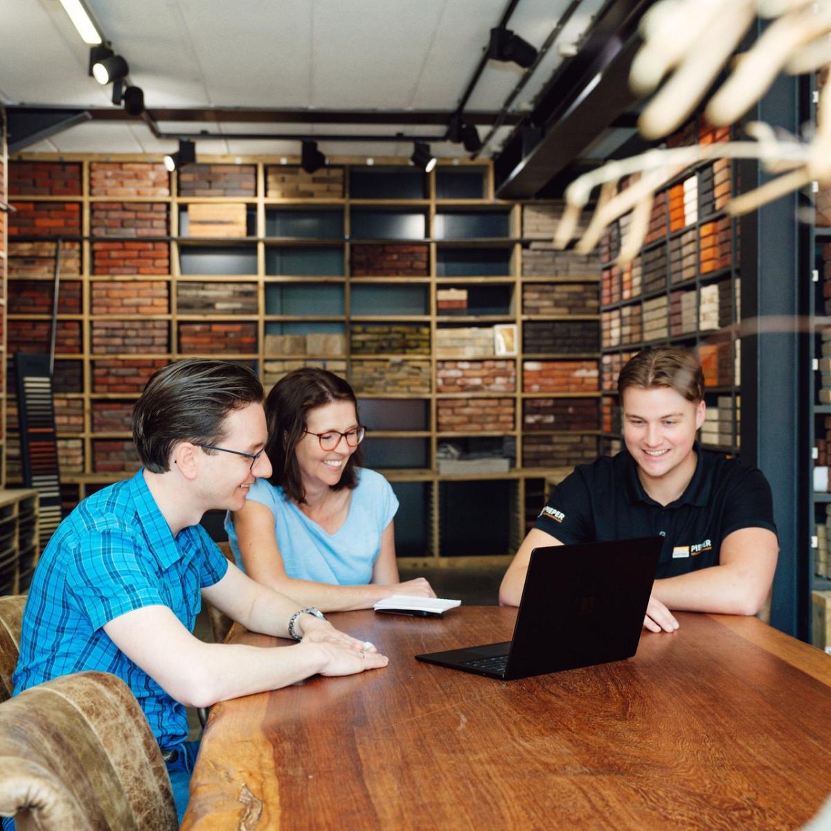 Drie mensen overleggen lachend aan een houten tafel in een showroom met een uitgebreide collectie baksteenstalen, terwijl een laptop openstaat.
