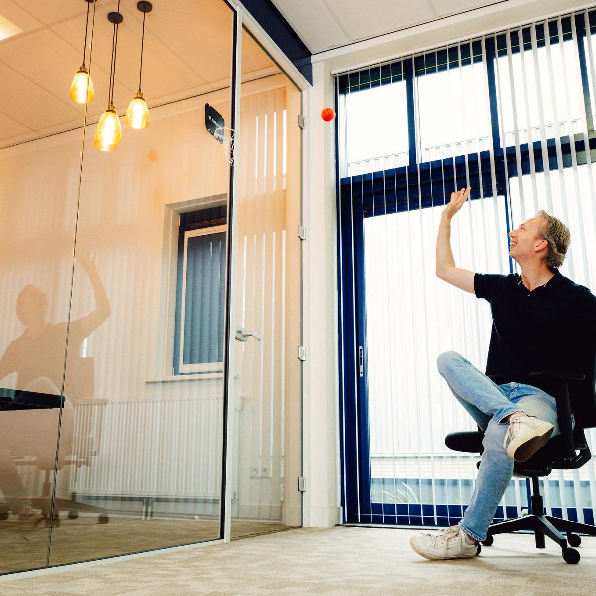 Man zit ontspannen op een bureaustoel in een kantoorruimte en gooit een rood balletje omhoog naast een glazen vergaderruimte.
