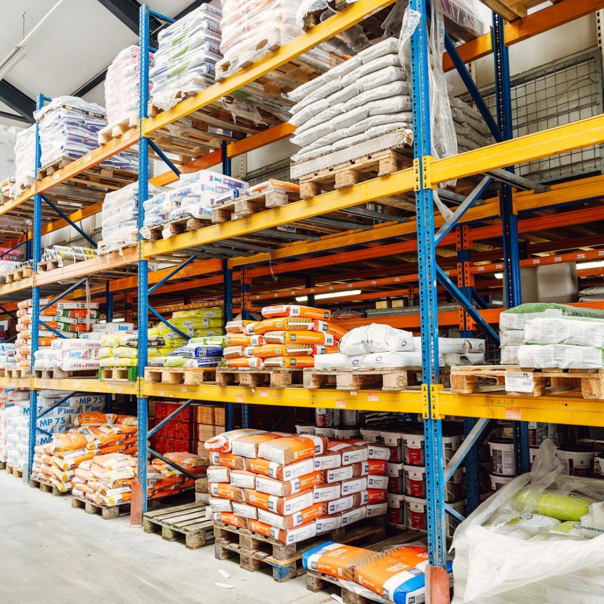 Opslagruimte met hoge stellingen gevuld met bouwmaterialen zoals cementzakken, isolatiemateriaal en verpakte producten, georganiseerd op pallets.