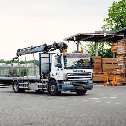 Vrachtwagen met kraan en heftruck staan op een bedrijfsterrein naast stellages met houten balken.