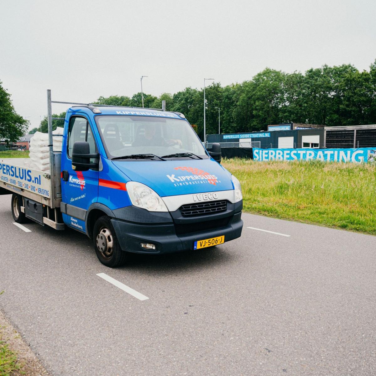 Bestelwagen op weg met lading, groene bomenrij en bewolkte hemel op achtergrond.