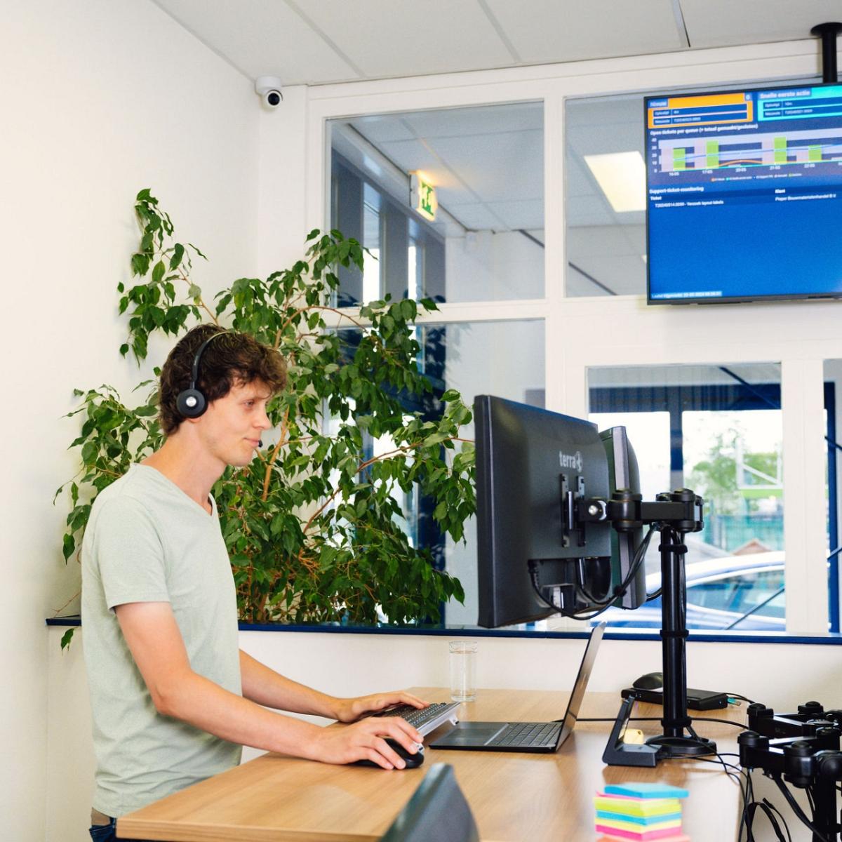 Een man staand aan een bureau met een koptelefoon op kijkt naar monitor, met boven hem een beeldscherm met gegevens in kantooromgeving.