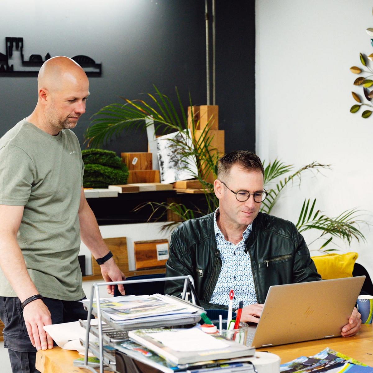 Een kantooromgeving met twee mannen kijkend op een laptop met op het bureau kantoorspullen en een monitor met informatie.