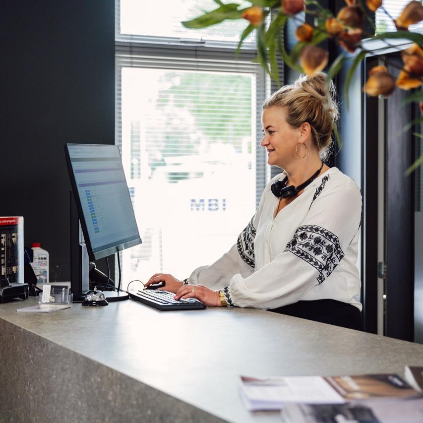 Vrouw staat achter een balie en kijkt op een beeldscherm met informatie en houdt handen op muis en toetsenbord in een werkomgeving.