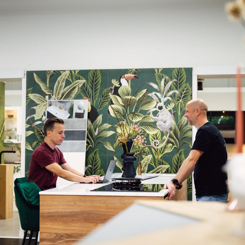 Twee mannen staan aan een keukenblok in een winkelomgeving met bloemdecoratie op de muur.