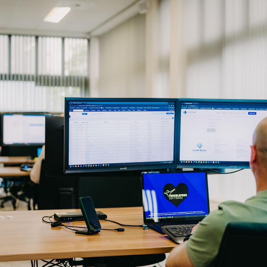 Twee monitors naast elkaar met informatie op een bureau waaraan iemand zit in een kantooromgeving.