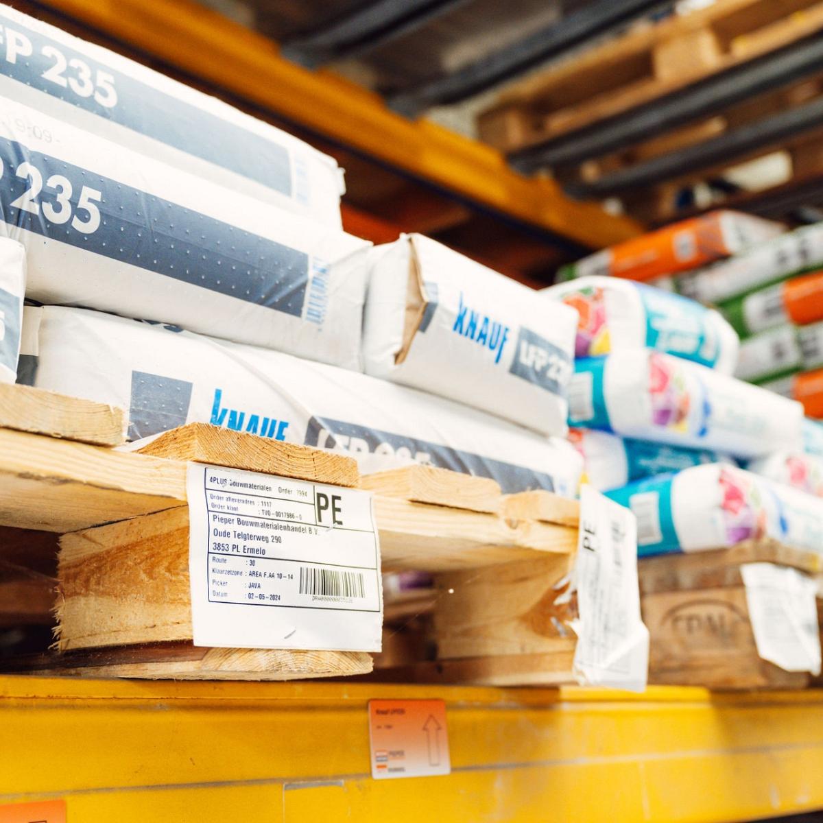 Close-up van houten pallets met daarop gestapelde bouwmaterialen, waaronder verpakkingen van Knauf LFP 235, voorzien van labels met productinformatie.