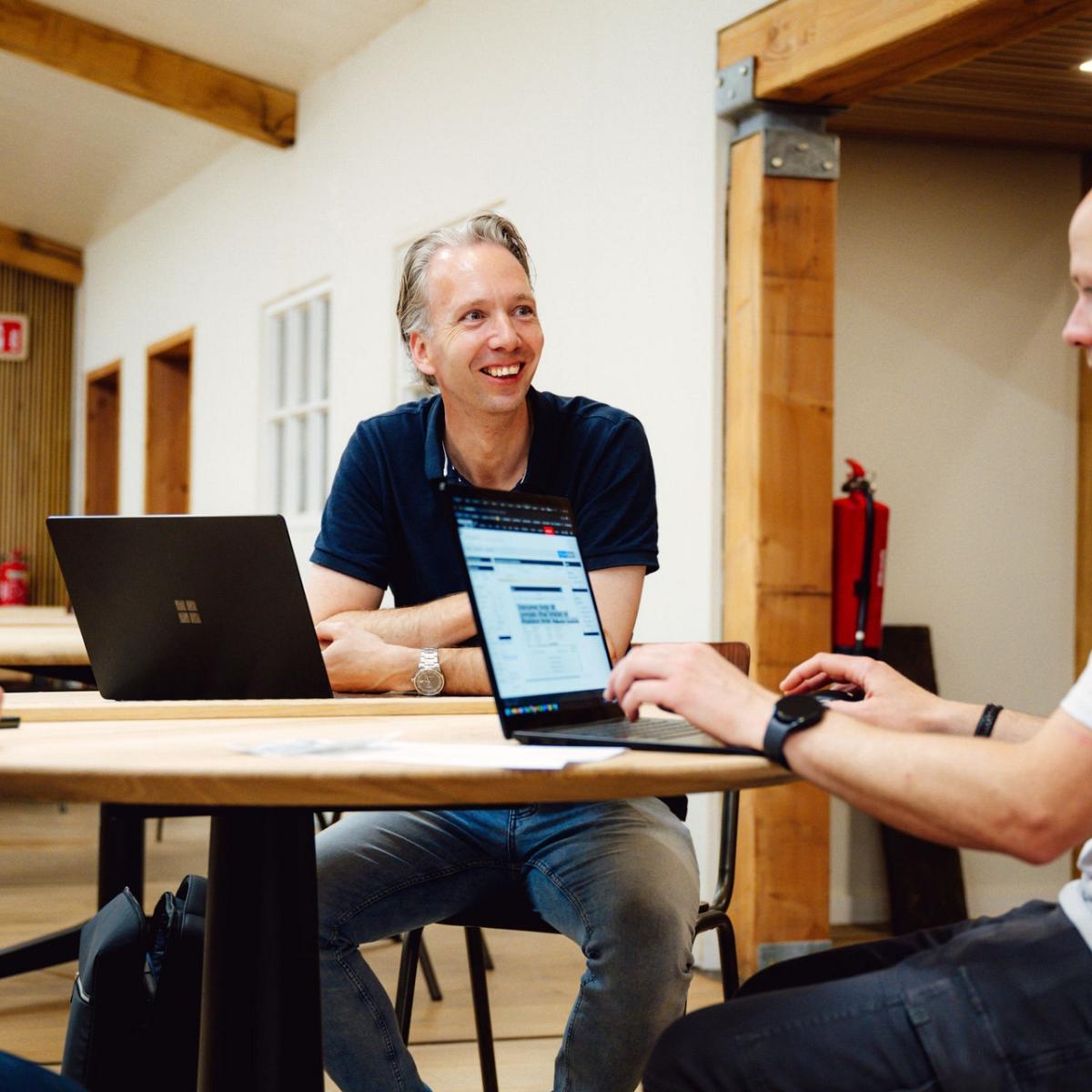 Drie mannen overleggen aan een houten tafel in een ruimte met houten accenten, waarbij een laptop openstaat met de website van Smart Trade op het scherm.