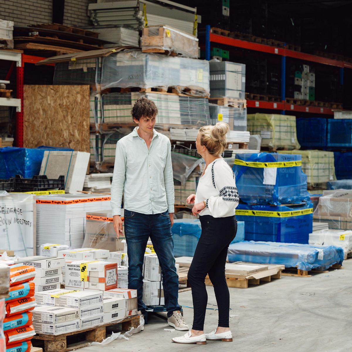 Twee mensen in gesprek tussen gestapelde pallets met bouwmaterialen in een magazijn, omringd door verpakkingen met tegellijm, isolatie en cementproducten.