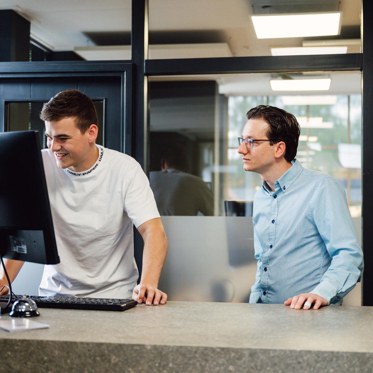 Twee mannen staan bij een balie en kijken samen naar een computerscherm, in een modern ingerichte ruimte met glazen wanden en strakke verlichting.