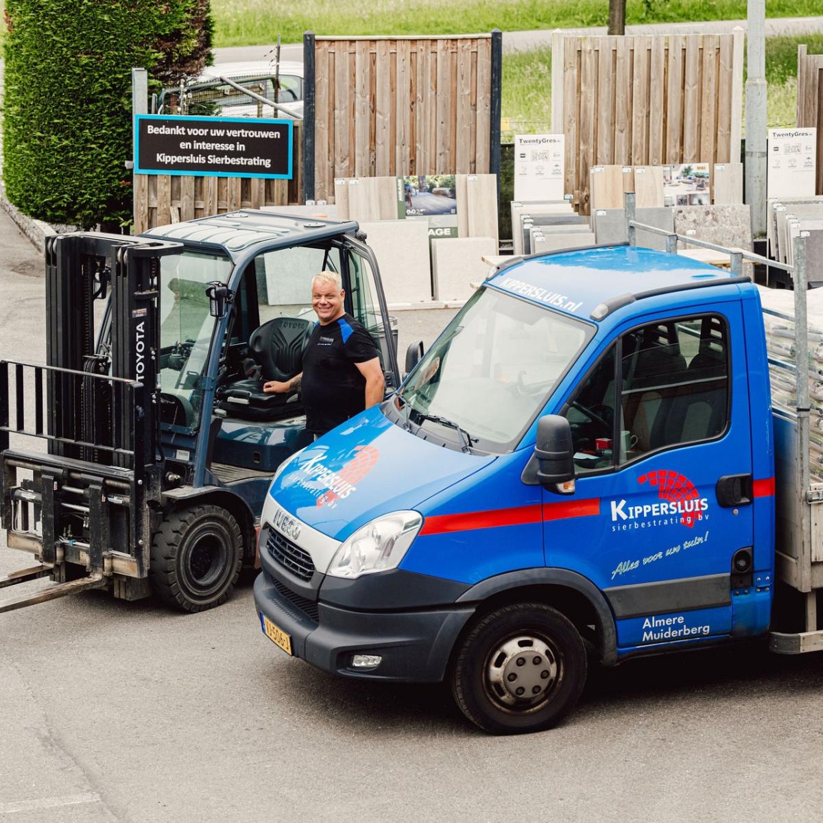 Een blauwe bestelwagen van Kippersluis Materialen met logo, naast een vorkheftruck bij een opslagplaats.