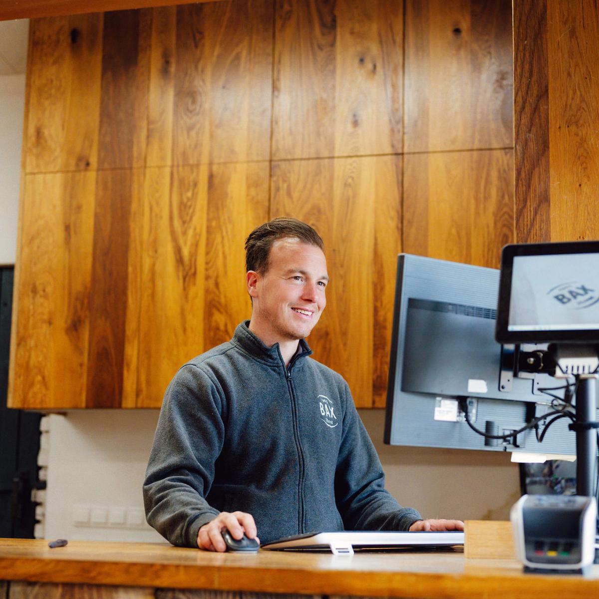 Medewerker achter een houten balie werkt met een computerscherm en muis, gekleed in een fleece trui met Bax-logo, in een interieur met houten wandbekleding.