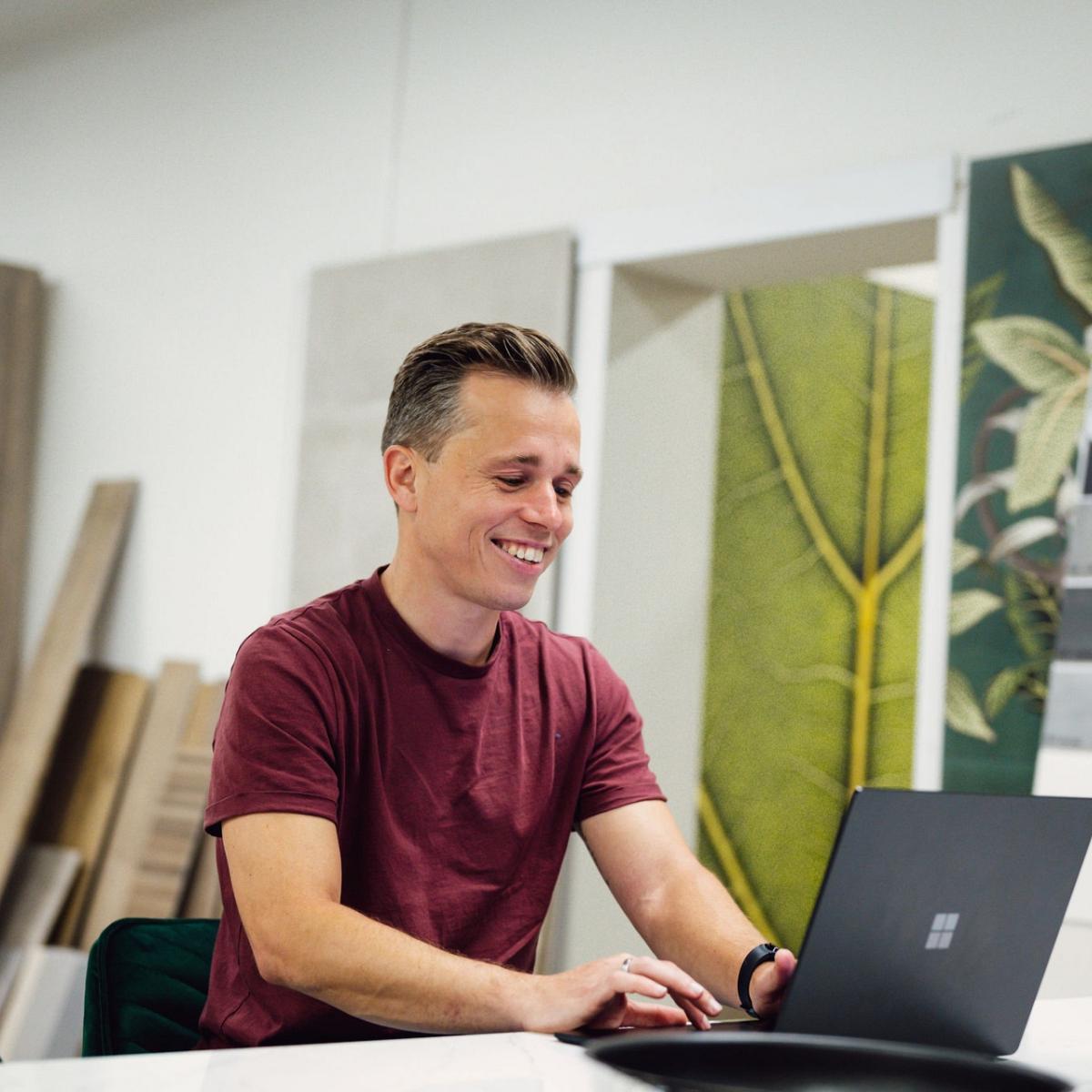 Man in een rood T-shirt werkt glimlachend op een laptop in een interieurshowroom, omringd door staaltjes van hout, tegels en decoratieve panelen.