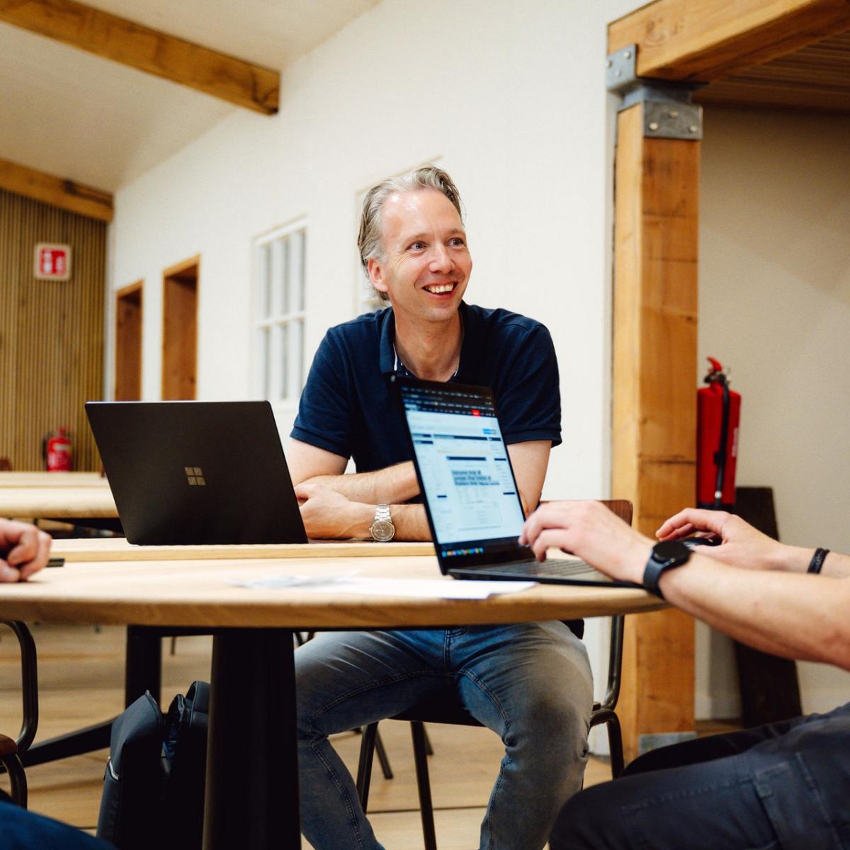 Drie mannen overleggen aan een houten tafel in een ruimte met houten accenten, waarbij een laptop openstaat met de website van Smart Trade op het scherm.