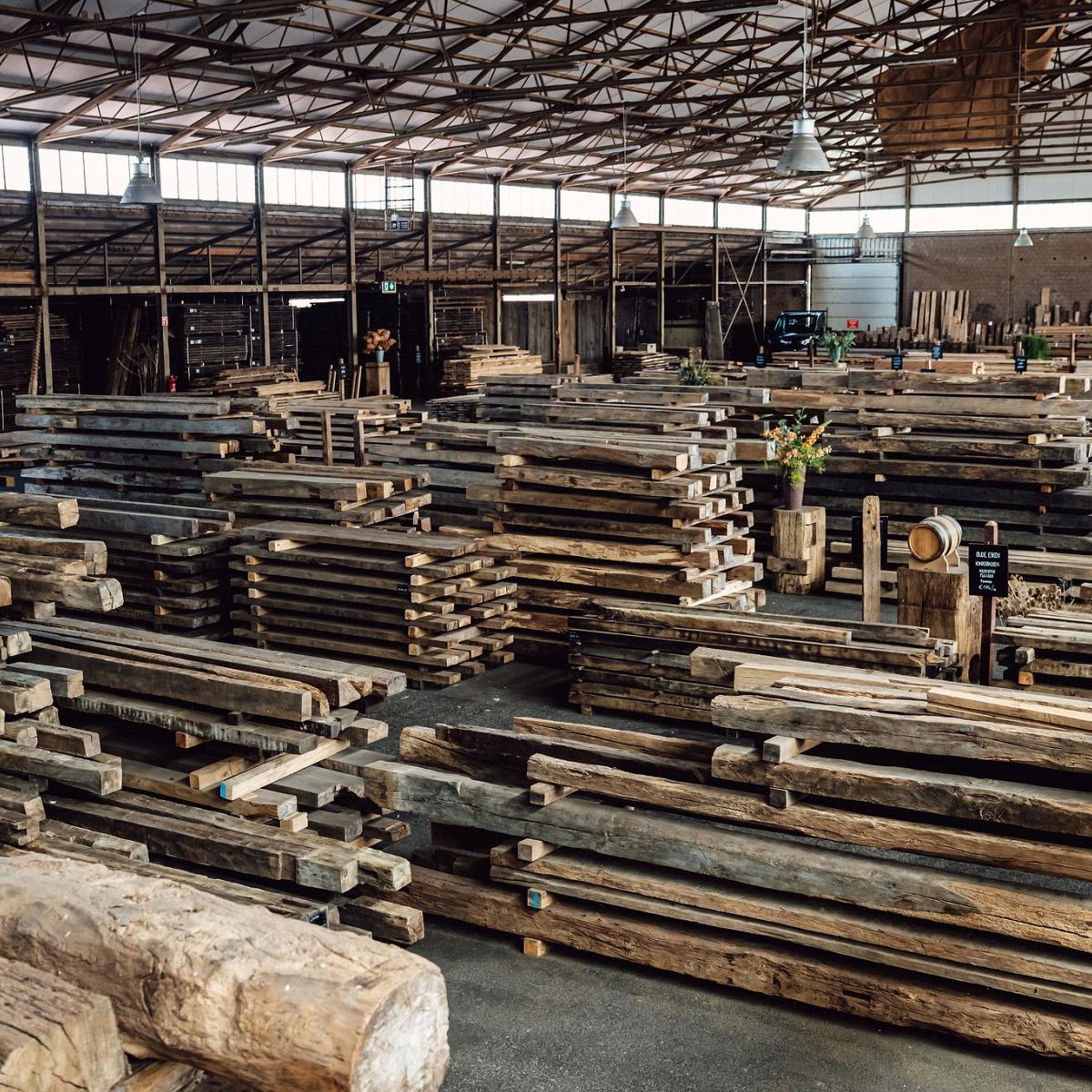 Grote overdekte houtloods met stapels ruw en gebruikt hout, gesorteerd op lengte en soort, klaar voor hergebruik of verkoop.